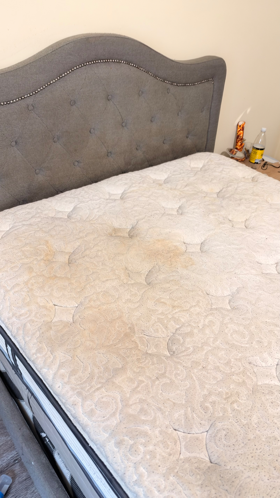 White mattress beside a grey tufted headboard with yellowish marks before cleaning
