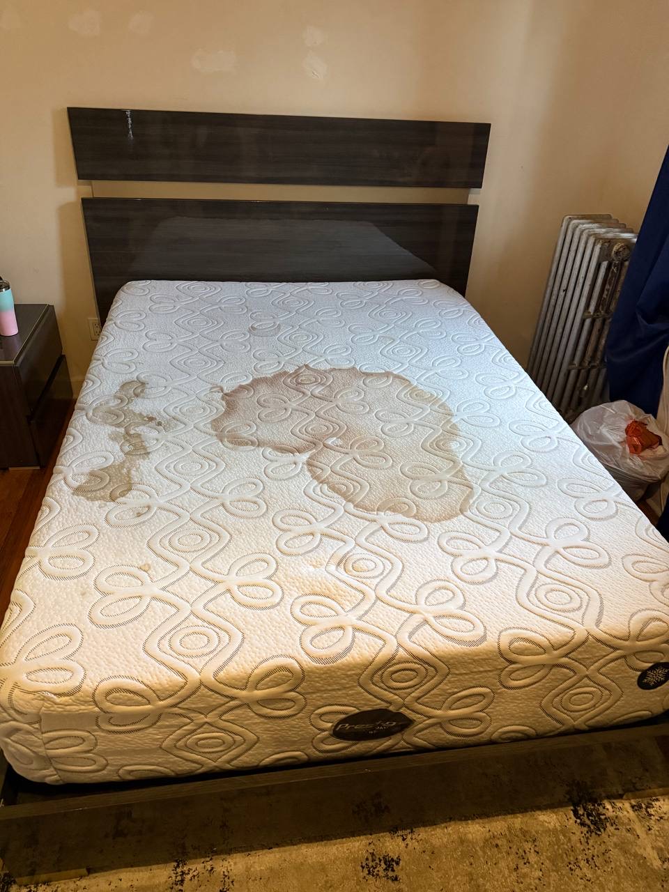 Patterned mattress beneath a wooden headboard with a large brown ring-shaped stain before cleaning